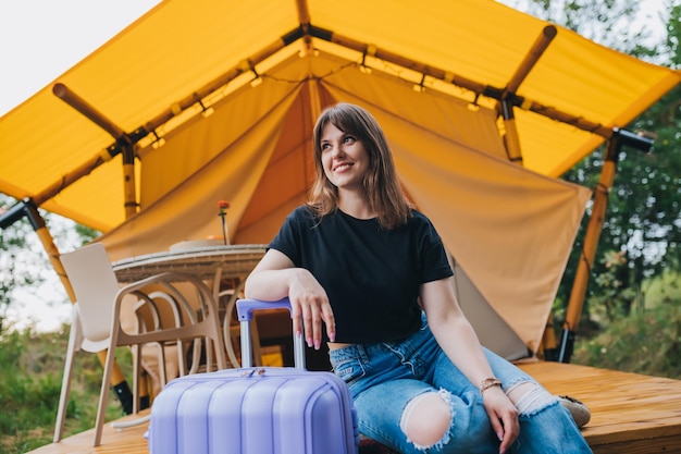 Happy woman traveler with luggage sitting on background of cozy glamping house Luxury camping tent for outdoor summer holiday and vacation Lifestyle concept