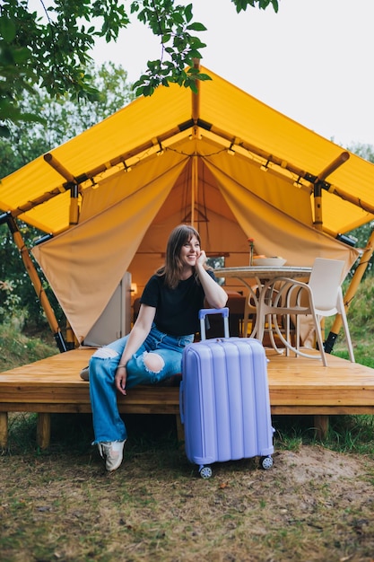 Felice donna viaggiatore con bagagli seduto sullo sfondo di un'accogliente casa glamping tenda da campeggio di lusso per vacanze estive all'aperto e vacanze concetto di stile di vita