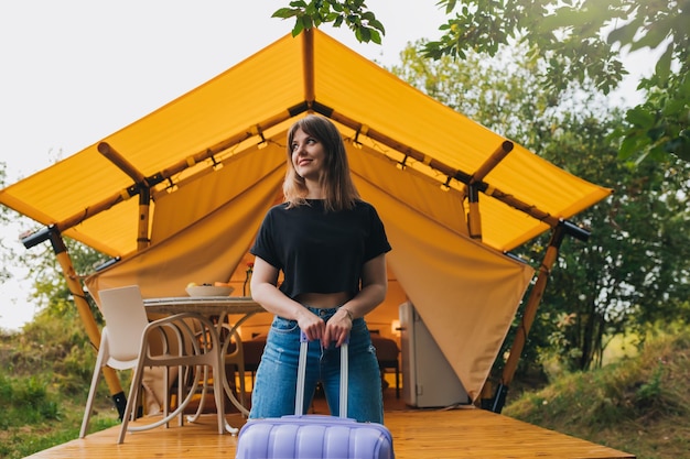 Happy woman traveler with luggage standing on background of cozy glamping house Luxury camping tent for outdoor summer holiday and vacation Lifestyle concept