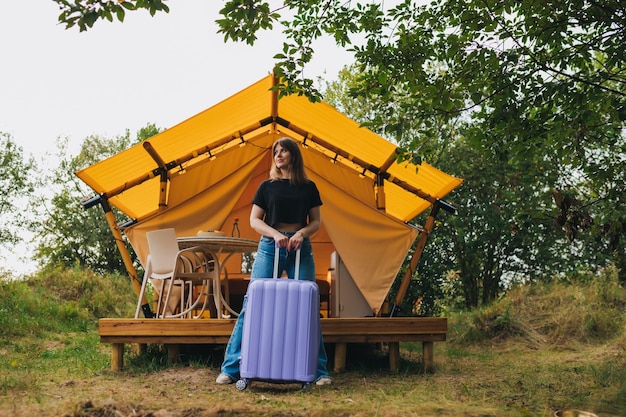 Felice donna viaggiatore con bagagli in piedi sullo sfondo di un'accogliente casa glamping tenda da campeggio di lusso per vacanze estive all'aperto e vacanze concetto di stile di vita