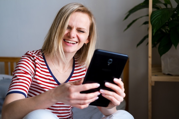 Happy woman using digital tablet for video call friends and parents, smiling girl sitting at home on bed fun greeting online by computer webcam making videocall