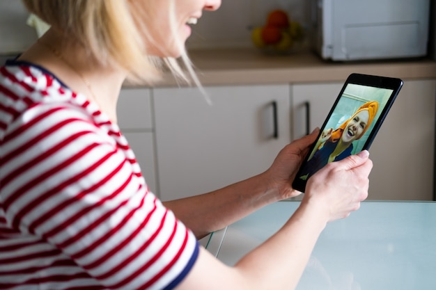 Donna felice che per mezzo della compressa digitale per gli amici e i genitori di videochiamata, ragazza sorridente che si siede a casa cucina saluto divertente online dalla webcam del computer che fa videocall