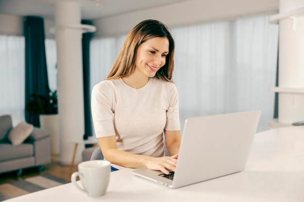 A happy woman using a laptop in kitchen at her home