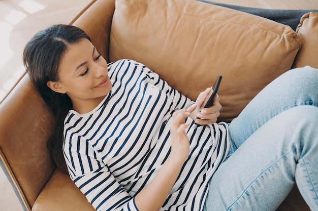 Happy woman using modern mobile apps on smartphone shopping in online store lying on sofa at home