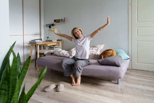 Happy woman waking up alone in morning at home sitting on bed with dog stretching body muscles