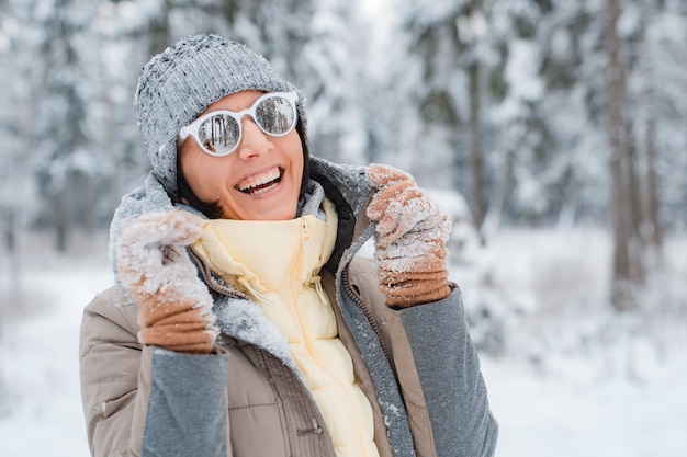 雪の降る冬の日に屋外を歩く幸せな女性。サングラスをかけた女性の幸せな陽気な服を着たファッショナブルな灰色は、冬の雪に覆われた森で喜ぶ