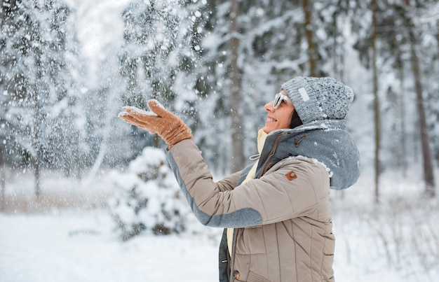 Happy woman walking outdoors on a snowy winter day. Female model dressed fashionable grey wearing sunglasses blowing snow having fun, catching snow with your hands