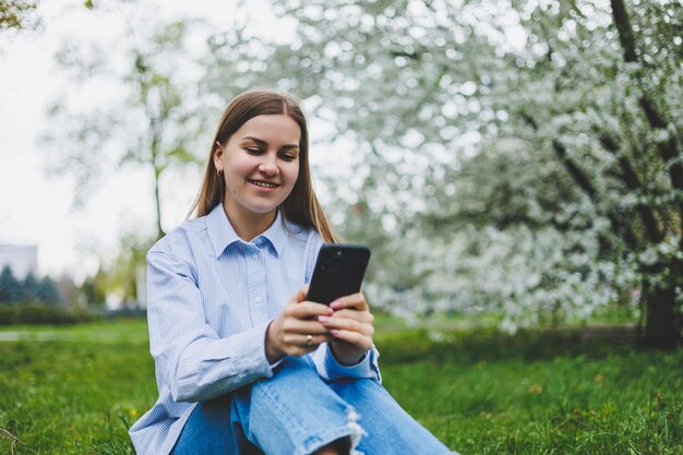 도시 거리에서 스마트 폰을 사용하여 걷는 행복한 여성 선글라스를 쓴 비즈니스 여성은 꽃이 만발한 나무 아래 야외에서 휴대 전화를 사용합니다.
