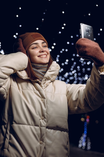 夜の街で携帯電話で自撮りをして楽しんでいる暖かい冬の服を着た幸せな女性