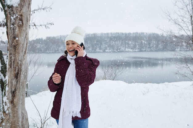 A happy woman in warm winter clothes talking on phone on the snow covered nature while falling snow on the lake background