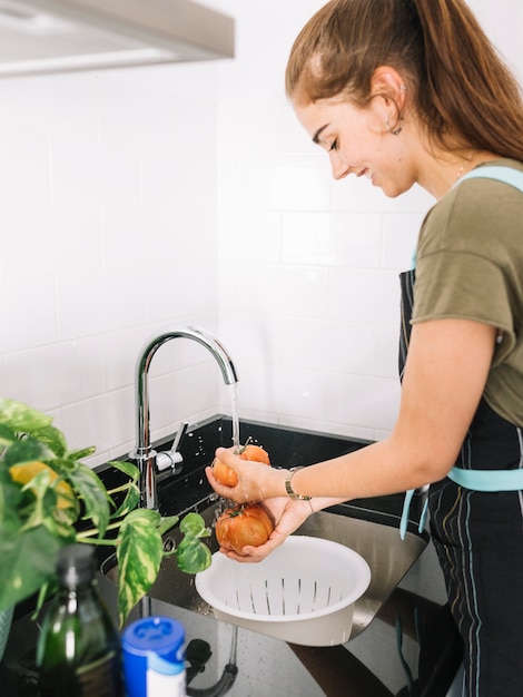 Photo happy woman washing red tomato in the kitchen sink