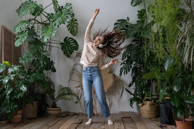 Happy woman wear wireless headphones listening to music dancing in home garden with tropical plants