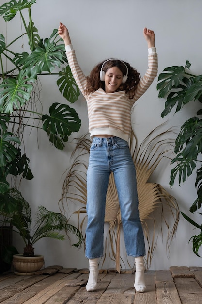 Happy woman wear wireless headphones listening to music dancing in home garden with tropical plants