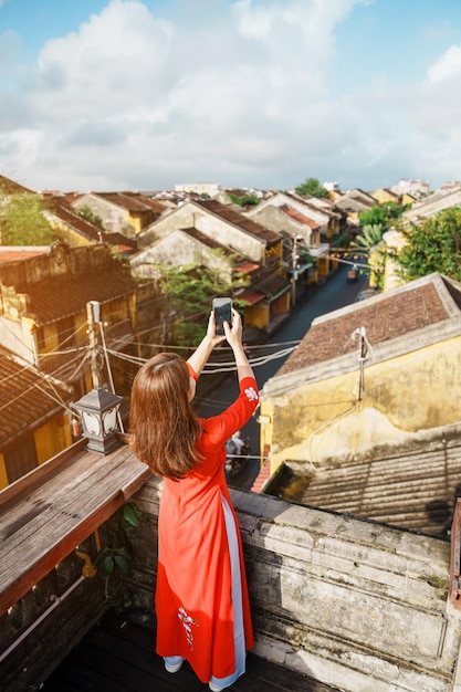 베트남 호이안 고대 도시 옥상에서 휴대폰으로 사진을 찍는 아오자이 베트남 드레스를 입은 행복한 여성과 동남아 여행 개념