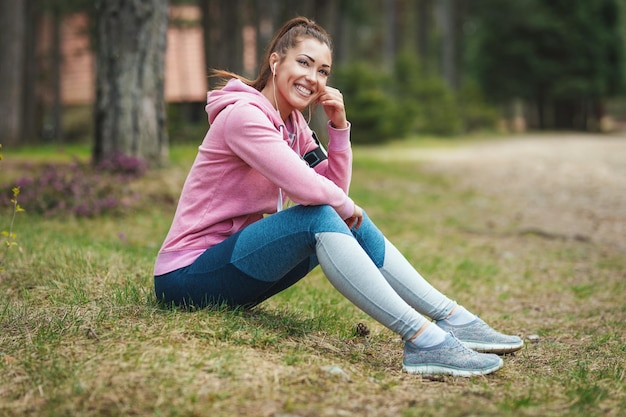 Happy woman, wearing sportswear, is sitting on the ground and, listening music in a break of exercises in a sunny forest.