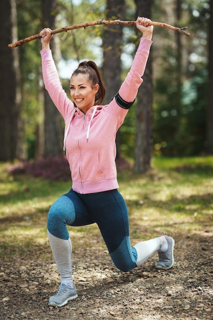 Photo happy woman, wearing sportswear, is stretching with branch in her hands, in a sunny forest trail, enjoying landscape among the trees.