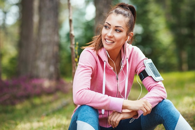 Happy woman, wearing sportswear, listening music in a break of exercises in a sunny forest, enjoying landscape among the trees.