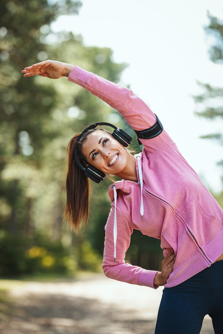 Photo happy woman, wearing sportswear, with headphones on her head is listening music and stretching in a sunny forest, enjoying landscape among the trees.