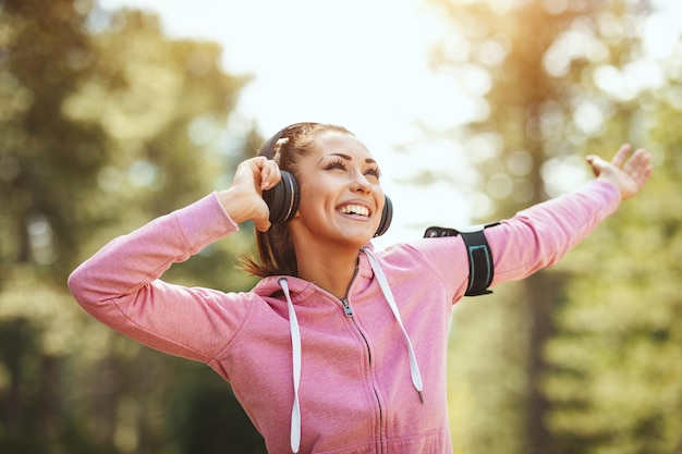 Happy woman, wearing sportswear, with headphones on her head is standing in a sunny forest, listening music and enjoying landscape among the trees.