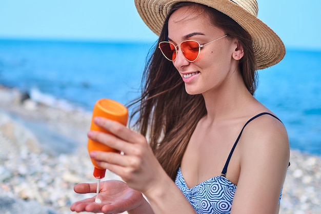 Donna felice che indossa il costume da bagno, cappello di paglia e occhiali da sole rosso brillante con la bottiglia di crema solare durante il sole al mare in tempo caldo e soleggiato in estate