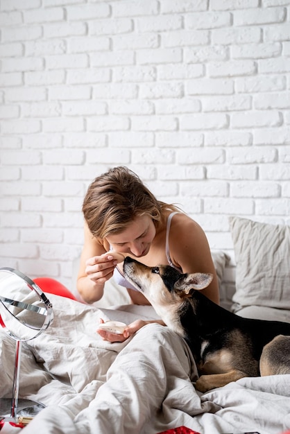 Happy woman wearing underwear doing routine skin care at home with beauty products Woman sitting on bed with her funny dog at home and applying cosmetics lifestyle pet care