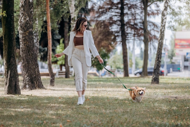 ウェルシュ・コーギー・ペンブローク犬と一緒に街の公園を歩く白いスーツとサングラスを着た幸せな女性