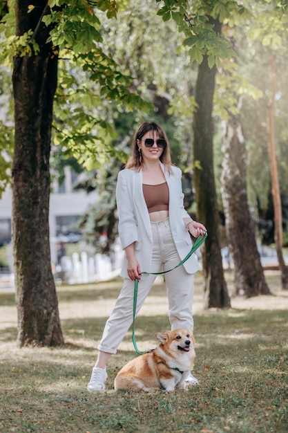 Happy Woman wearing white suit and sunglasses walking in city parkland with Welsh Corgi Pembroke dog