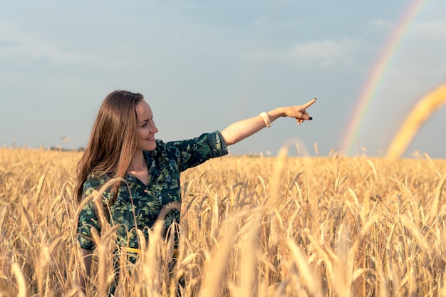 Happy woman in wheat field with Golden spikelets pointing her finger at rainbow