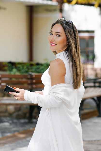 Happy woman in white clothes walks around the city holding a smartphone in her hand