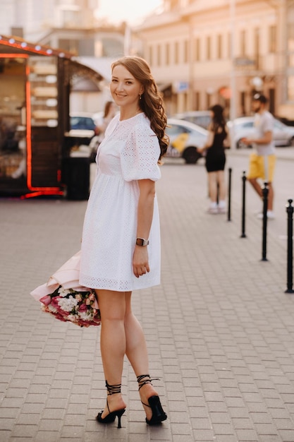 A happy woman in a white dress at sunset with a bouquet of flowers in the city