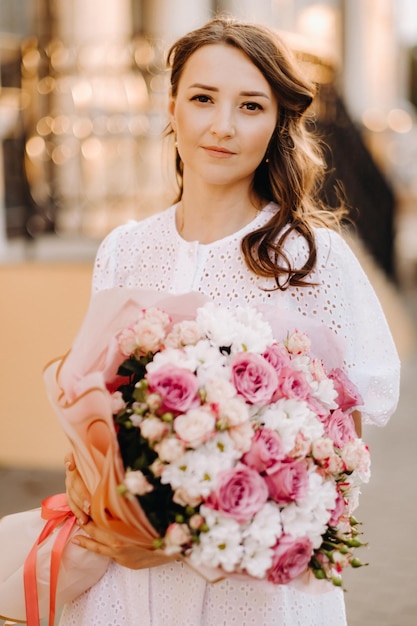 A happy woman in a white dress at sunset with a bouquet of flowers in the city