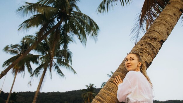 Happy woman in a white shirt on beach near stormy sea tilted palm tree Attractive girl with wet curly hair in sunglasses Female tourist walks has fun rest Camera follow spin rotate around model