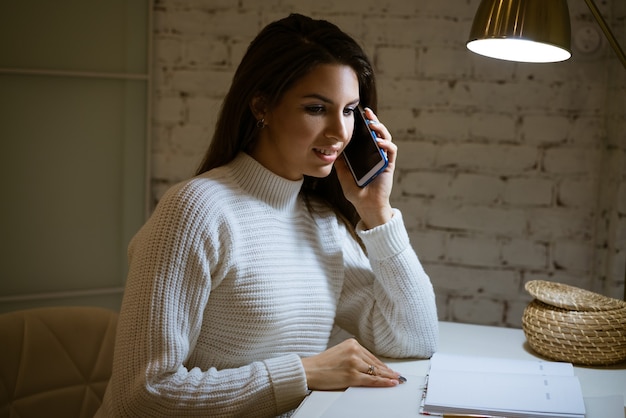 Happy woman in white sweater talking on the phone at the table