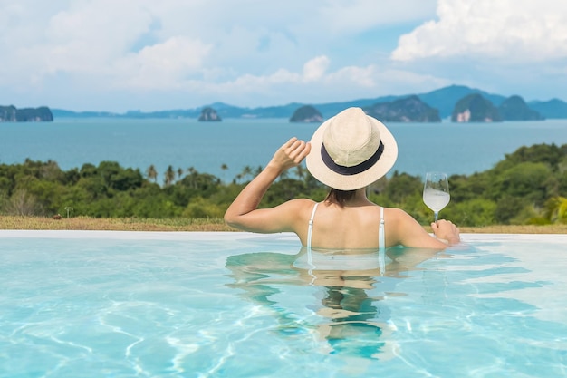 Happy woman in white swimsuit drinking wine in luxury swimming pool hotel against beautiful view young female with hat enjoy in tropical resort Relaxing summer travel holiday and vacation concept