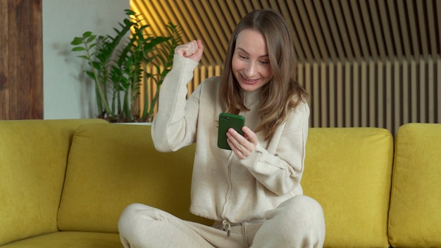 Happy woman winner holding smartphone reading message celebrating mobile victory, using cellphone sit on sofa at home.