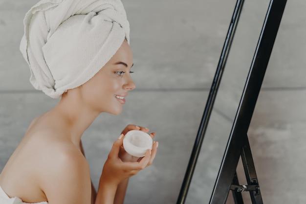 Happy woman with bare shoulders towel on head applies face cream admires reflection Selfcare