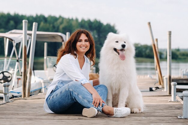 A happy woman with a big white dog lies on a pier near the sea at sunset