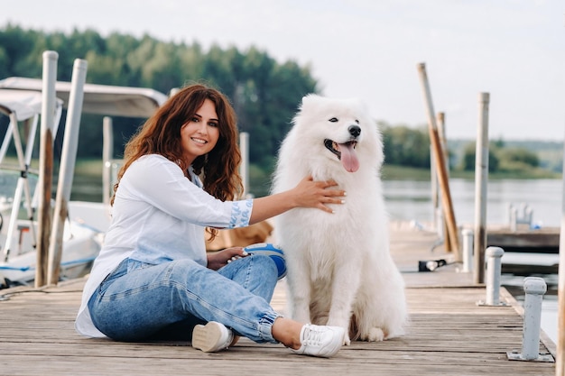 A happy woman with a big white dog lies on a pier near the sea at sunset