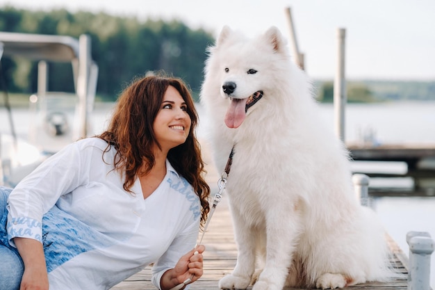 A happy woman with a big white dog lies on a pier near the sea at sunset