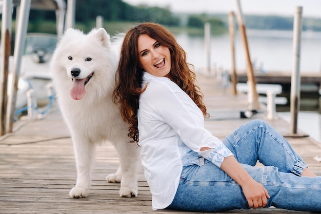 A happy woman with a big white dog sits on a pier by the sea at sunset.