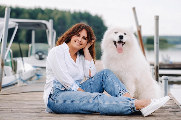 大きな白い犬と幸せな女性は日没時に海のそばの桟橋に座っています