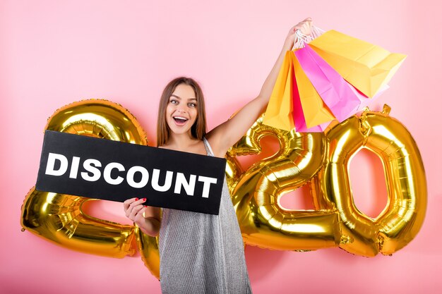 Happy woman with colorful shopping bags and copyspace discount sign with 2020 balloons isolated over pink