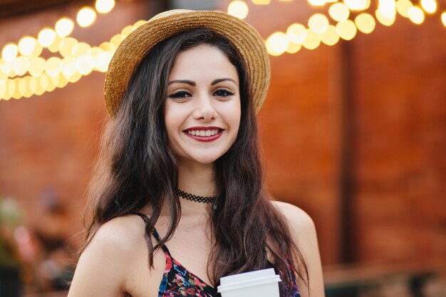 Happy woman with dark hair charming eyes and gentle smile wearing straw hat having summer vacations spending free time at terrace drinking takeaway coffe from paper cup enjoying her drink