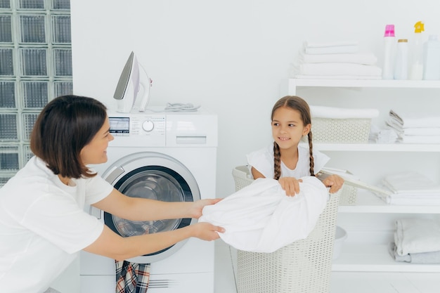 Happy woman with dark short hair pulls off laundry from basket\
happy child poses in it spend time in bathroom near washing machine\
and iron on top shelf with folded white towels laundry time