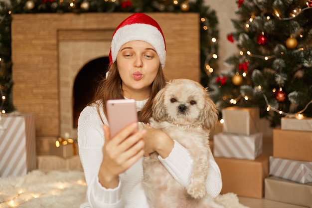 Happy woman with dog take selfie in christmas decoration, woman blowing kiss gesture to camera of smart phone, keeping lips rounded, wearing white casual jumper and santa claus hat.