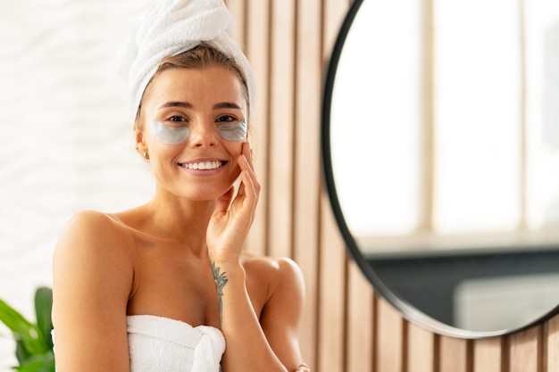 Happy woman with eye patches after shower wearing white towel standing in bathroom near mirror