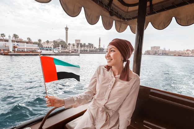 Happy woman with the flag of the United Arab Emirates sits in abra dhow boat during a cruise on the Dubai Creek Canal with scenic views of numerous skyscrapers and attractions