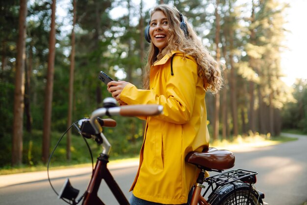 Счастливая женщина с наушниками и смартфоном ездит на велосипеде в солнечном парке. Активный образ жизни отдыхает.