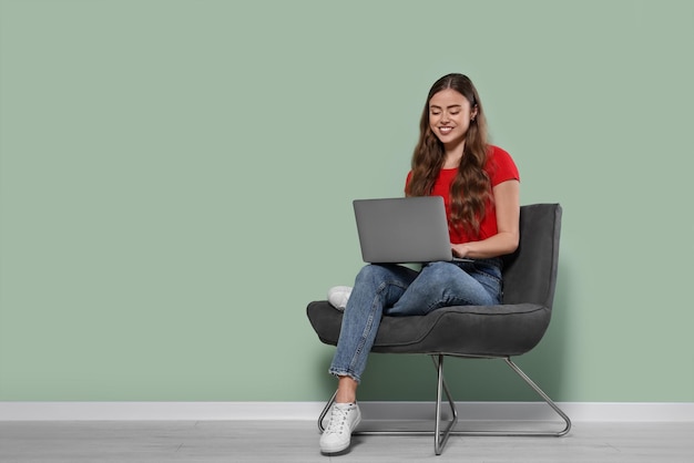 Happy woman with laptop sitting in armchair near pale green wall indoors Space for text