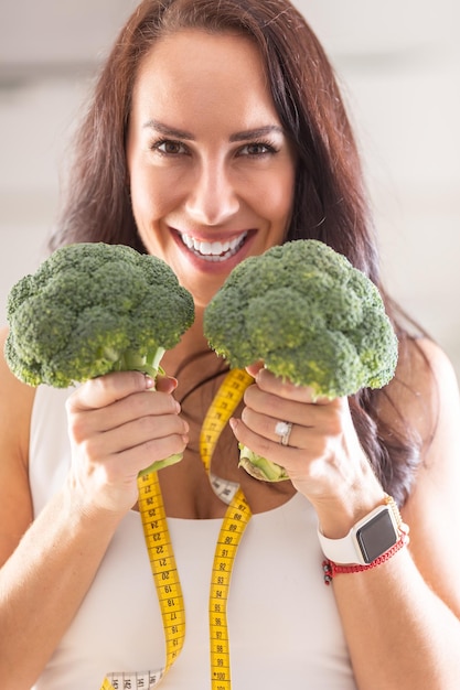 Happy woman with a measuring tape around her neck holds two broccolis in her hands smiling